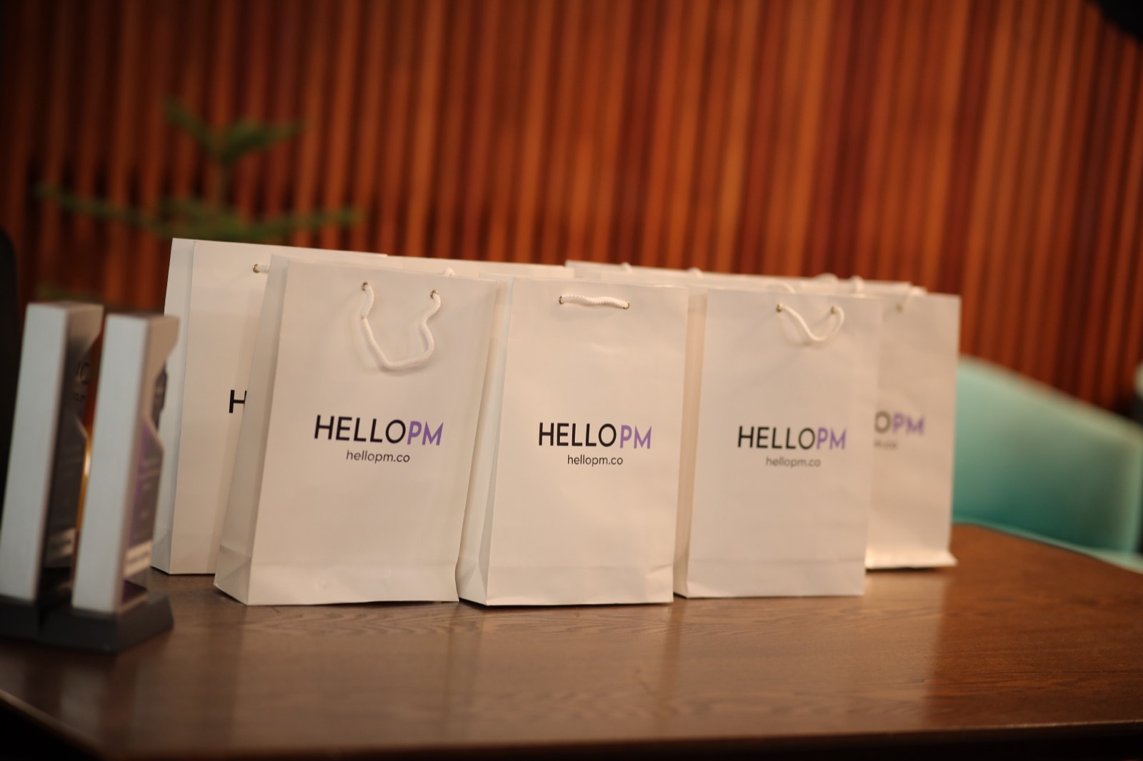 A row of white HelloPM branded gift bags lined up on a table beside event trophies