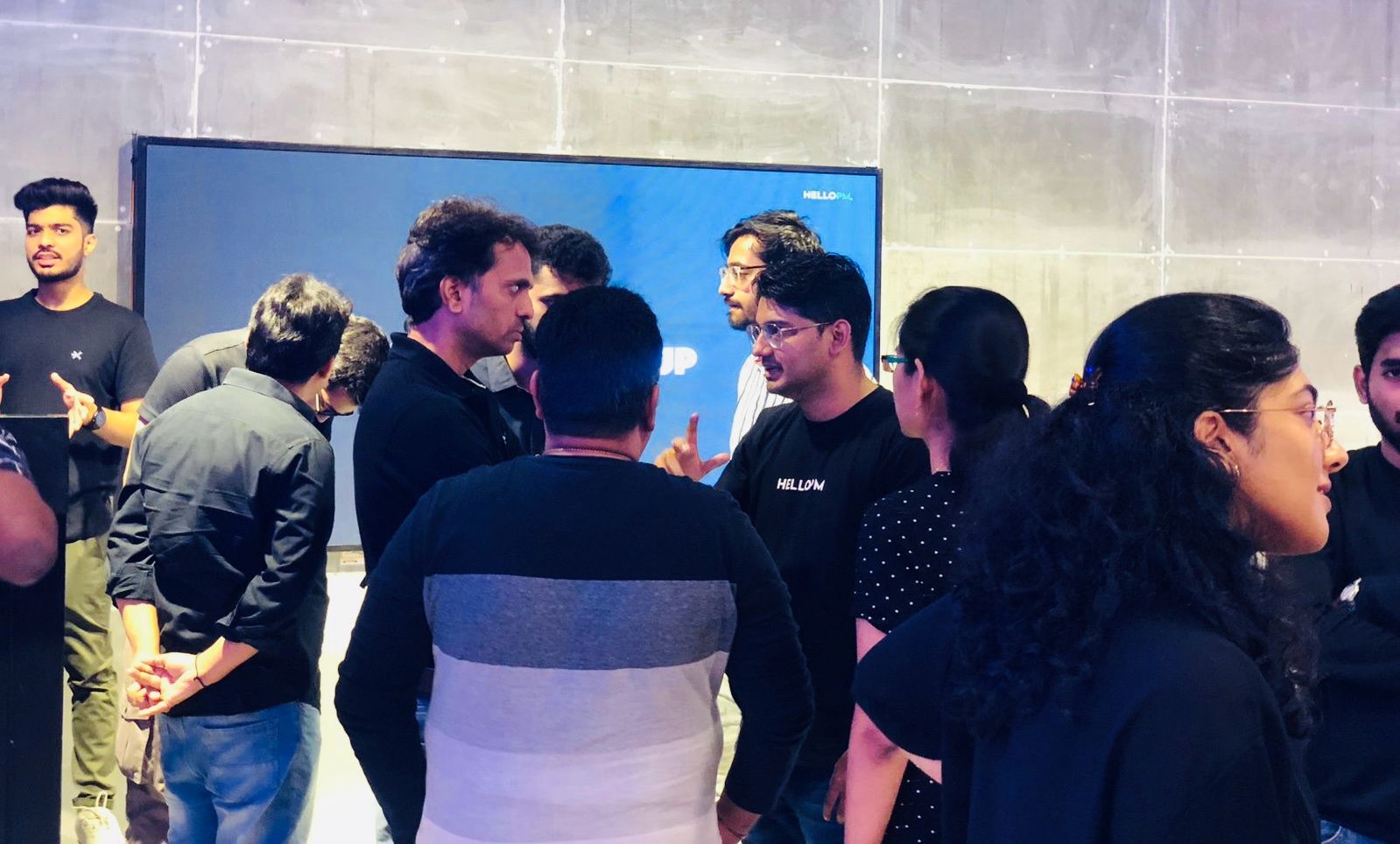 A cluster of HelloPM members networking in front of a HelloPM screen
